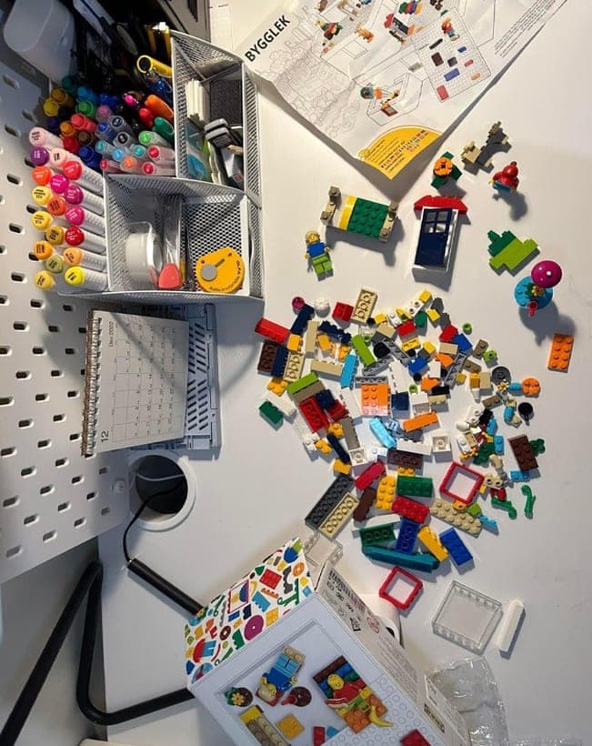 LEGO bricks, minifigures and building instructions spread across a white desk beside a pegboard of coloured markers