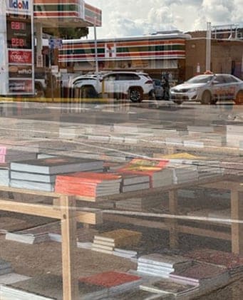 Books on display tables in a shop window, with a petrol station and parked cars reflected in the glass