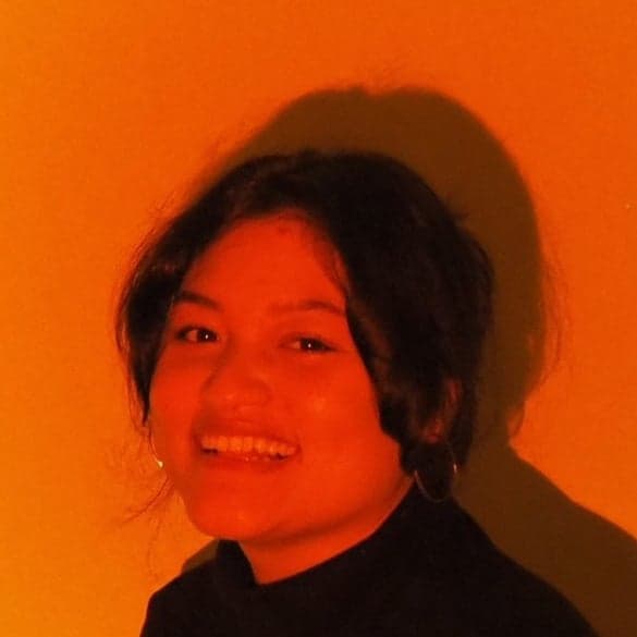 Portrait of Parnvard Amnuay smiling, lit in warm orange light against a plain wall